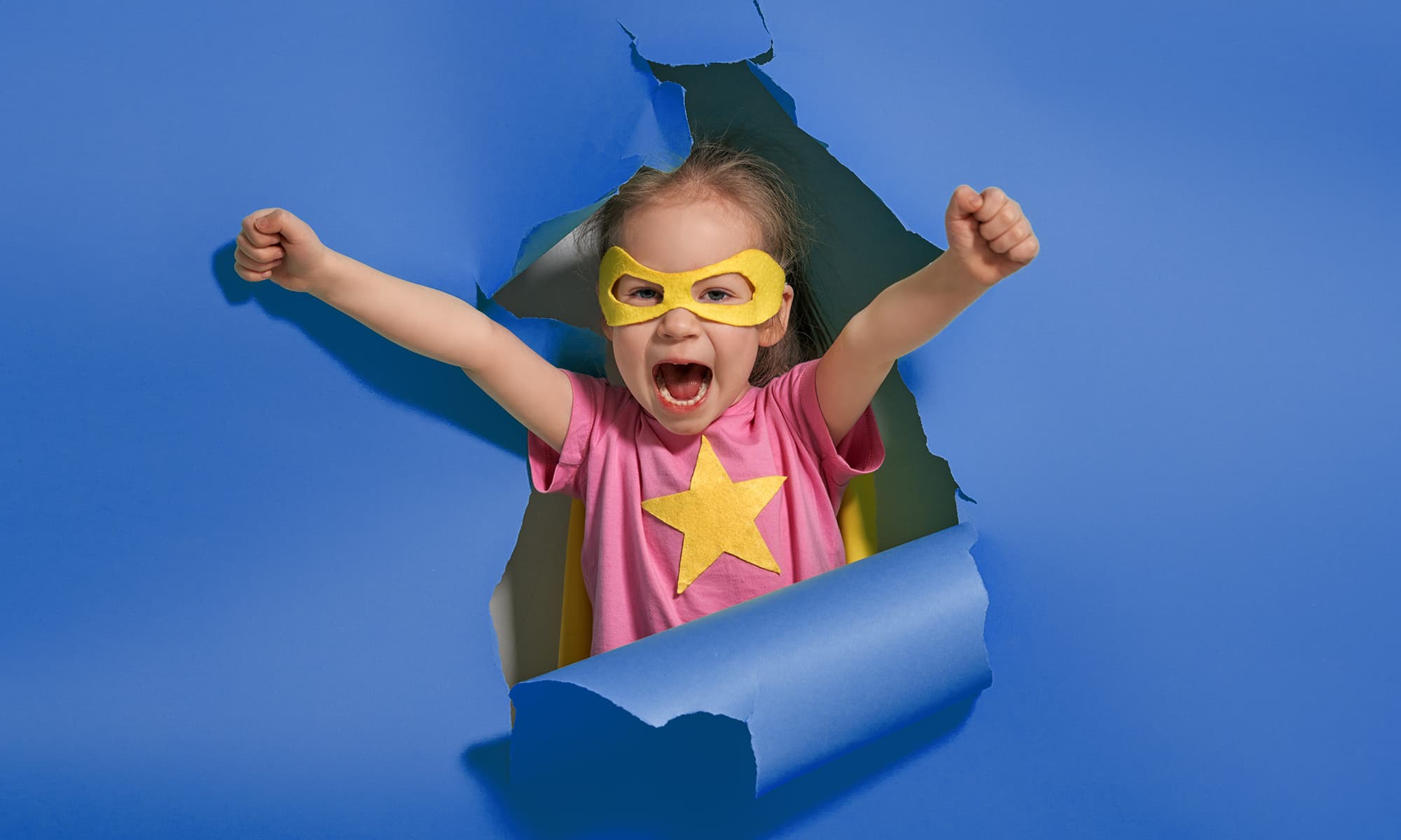 A joyful child in a yellow superhero mask and star, arms raised in the air