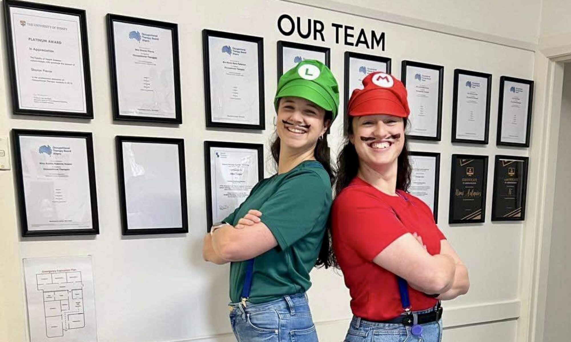Two OT for Kids therapists in front of the practice’s wall of framed AHPRA Occupational Therapy Board registrations and a University of Sydney award